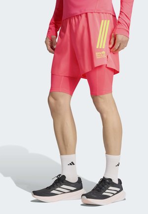 Hombre que lleva pantalones cortos de capas de color rosa brillante con texto "BMW Berlin Marathon", calcetines blancos Adidas y zapatillas de correr Adidas en blanco y negro.