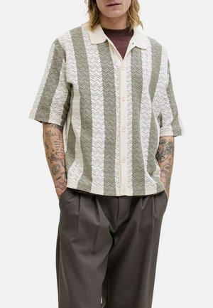 Mann iført en kortermet, knappet skjorte med krage og brede vertikale beige og olivengrønne striper, kombinert med løse, mørke bukser og synlige tatoverte armer.