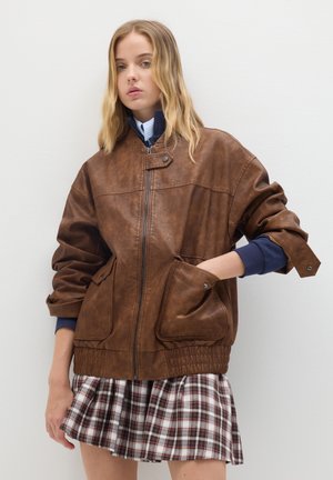 Jonge vrouw met lang blond haar, gekleed in een brown bikerjack over een marineblauwe sweatshirt en een geplisseerde geruite rok, met haar hand in haar zak.