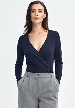 Granatowy bodysuit z długim rękawem, z głębokim dekoltem w serek i zwiewnym krojem, zestawiony z jasnoszarymi spodniami z wysokim stanem, z przodem w kant i guzikiem.