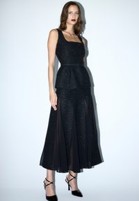 Schwarzes Peplum-Kleid mit strukturiertem Stoff, quadratischem Ausschnitt und transparenten Rockpaneelen. Kombiniert mit schwarzen Riemchen-Heels.