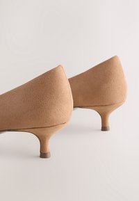 Chaussures à talons hauts en suède beige avec un embout arrondi et un talon fin. La texture est lisse avec une finition mate et des détails de couture minimalistes.