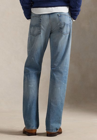 Personne portant un jean en denim bleu, des chaussures marron, une chemise blanche et un pull bleu foncé, debout contre un fond uni, vue de dos.