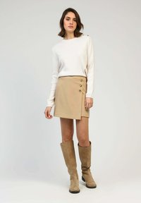 Jupe enveloppante beige avec trois boutons décoratifs, associée à une chemise blanche à manches longues et des bottes en daim beige jusqu'aux genoux. Texture de tissu lisse.