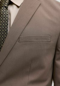 Detailní záběr na béžové sako s klopami s výřezem, náprsní kapsou, béžovou košilí a černo-béžovou kostkovanou kravatou.