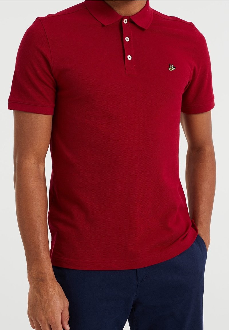 Röd pikétröja gjord av texturerad tyg, med klassisk krage och treknapps knäppning, prydd med en liten grön logotyp på bröstet.