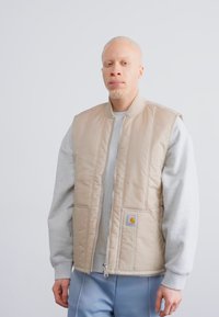 Vest bej cu umplutură, cu închidere cu fermoar, hanorac gri dedesubt și patch cu logo pe partea stângă. Material neted, croială lejeră, buzunare laterale vizibile.