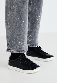 Zapatillas de cuero negro con suelas de goma blanca y detalles texturizados. Combinadas con unos jeans de mezclilla gris decorados con puntos plateados embellecidos.
