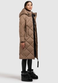 Beige vuorattu pitkä takki, jossa on huppu, vetoketjullinen etuosa, sivutaskut ja hieman kiiltävä pinta, yhdistettynä mustiin talvikenkien kanssa.