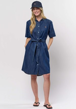 Žena se usmívá, má na sobě modré džínové šaty na knoflíky s páskem, odpovídající čepici a černé žabky, stojí s rukama v kapse.