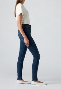 Lehké bílé tričko, tmavě modré skinny džínsy s jemnými švy a bílé ploché boty, pózující v profilu na neutrálním pozadí.