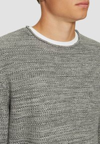 Grijze gebreide trui met een textuurpatroon, voorzien van een ronde halslijn en een gelaagd wit shirt dat eronder zichtbaar is.