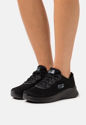 Pieds portant des chaussures de sport Skechers noires à lacets, avec des empiècements en mesh et des semelles rembourrées sur un fond blanc.