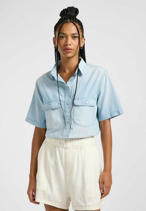 Camisa de botón de manga corta azul claro con dos bolsillos en el pecho y cuello, combinada con pantalones cortos de tiro alto color crema con detalles de costura visibles.