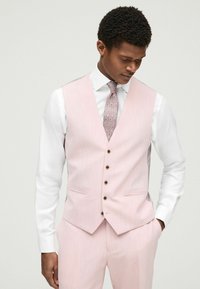 Gilet rosa chiaro con effetto testurizzato, dotato di bottoni marroni e due tasche frontali. Indossato sopra una camicia bianca e una cravatta a fantasia.