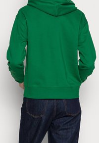 Personne portant un sweat à capuche vert uni et un jean bleu foncé, vue de dos sur un fond uni.