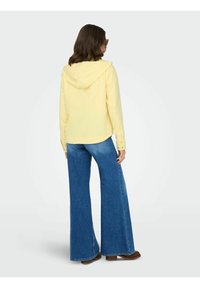 Femme aux cheveux bruns mi-longs portant une veste à capuche jaune pâle, un jean large bleu et des chaussures marron, de dos.