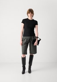 Svart teksturert kortermet topp, olivenfargede shorts og knelange svarte støvler. Holder en liten svart håndveske. Minimalistisk styling.