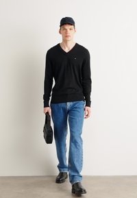 Pulover negru tricotat cu decolteu în V, blugi blue straight-leg, geantă neagră și pantofi negri, purtat cu o pălărie închisă. Outfit minimalist, casual.