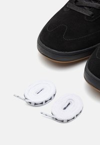 Czarne zamszowe trampki z gumowymi podeszwami, z czarnymi sznurowadłami, obok dwóch zwiniętych białych sznurowadeł z czarnymi akcentami.