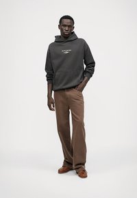 Hanorac gri închis cu buzunar tip cangur și logo, asortat cu pantaloni maro cu croială largă și mocasini maro. Aspect simplu și casual.