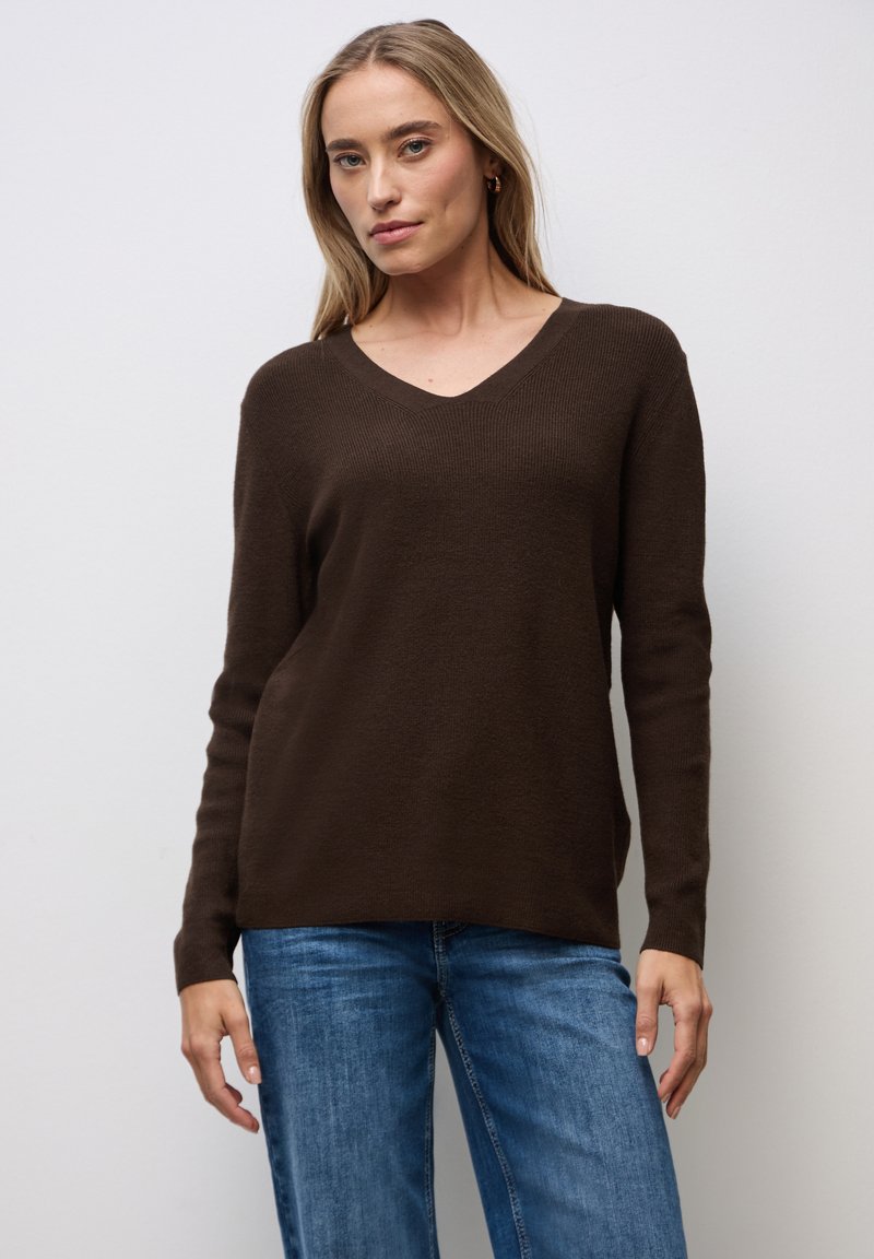 Brązowy sweter w serek z miękkiego, teksturowanego dzianiny z długimi rękawami, w połączeniu z niebieskimi jeansami o prostym kroju. Prosty design, bez wzorów czy akcentów.