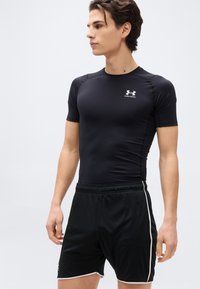 Νέος άνδρας φορώντας μαύρο συμπιεστικό μπλουζάκι Under Armour και μαύρο αθλητικό σορτς με λευκή επένδυση, στέκεται μπροστά σε απλό λευκό φόντο.