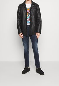 Svart läderjacka med krage, knappstängning, bärs över en färgglad grafisk t-shirt, i kombination med mörkblå jeans och svarta skor.