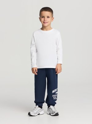 Maglietta bianca a maniche lunghe, pantaloni della tuta blu navy con testo bianco e scarpe da ginnastica argentate. Design semplice, vestibilità comoda e polsini a coste.