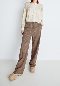 Donna che indossa una blusa color crema e pantaloni lunghi a quadri marroni con scarpe beige a suola spessa, in piedi su uno sfondo bianco.