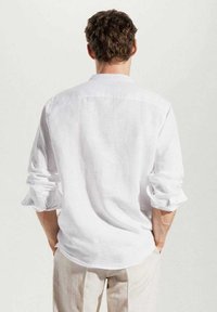 Uomo con capelli ricci che indossa una camicia bianca a maniche lunghe con le maniche arrotolate e pantaloni beige, in piedi di spalle con le mani nelle tasche.