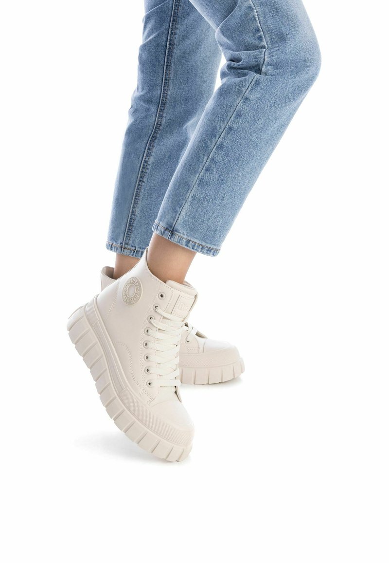 Zapatillas altas blancas con suela gruesa y texturizada, con un logotipo redondo, cordones en la parte delantera y material superior liso. Combinadas con jeans de mezclilla azules.