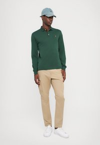 Polo de manga larga verde oscuro con cuello, gorra de denim claro, pantalones cargo beige y zapatillas blancas. Tejido suave, diseño ajustado.