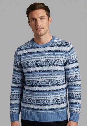 Uomo che indossa un maglione lavorato a maglia blu e bianco con motivi di fiocchi di neve e disegni geometrici, in piedi davanti a uno sfondo grigio uniforme.