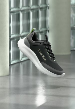 Baskets Calvin Klein noires et grises avec semelle blanche et accents verts flottant à l'intérieur contre un mur en briques de verre et un sol poli.