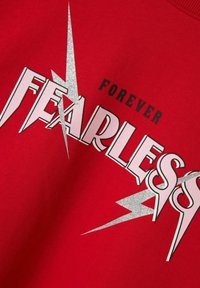 Felpa rossa con la parola "FEARLESS" in lettere grasse bianche e rosa, con accenti di fulmini argentati e la parola "FOREVER" sopra.