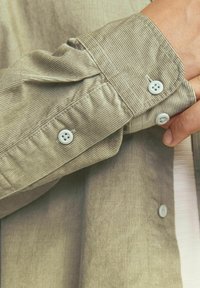 Cămașă din fustă khaki cu manșete cu nasturi. Prezintă nasturi albi și o textură cu nervuri verticale, având un aspect moale și subtil strălucitor.