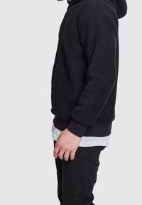 Felpa nera in pile con superficie testurizzata, polsini a coste e orlo a bande, sovrapposta a una camicia bianca per creare contrasto.