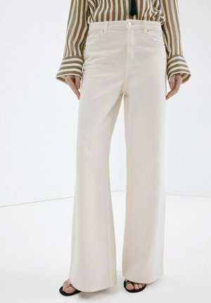 Mujer con pantalones crema de talle alto y pierna ancha, combinados con una camiseta de manga larga a rayas beige y blanca, y sandalias negras con punta abierta.