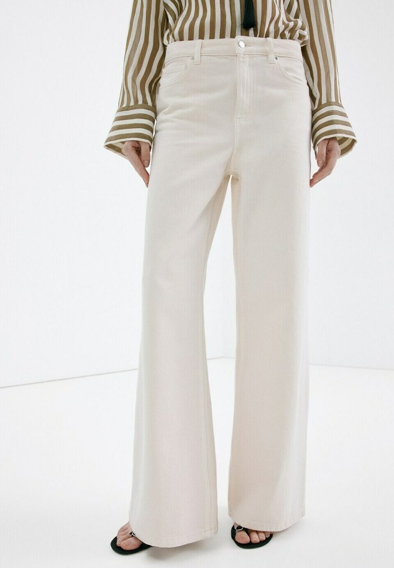 Femme portant un pantalon crème à taille haute et jambes larges, assorti d'un chemisier à manches longues beige et blanc à rayures, ainsi que des sandales noires à bouts ouverts.