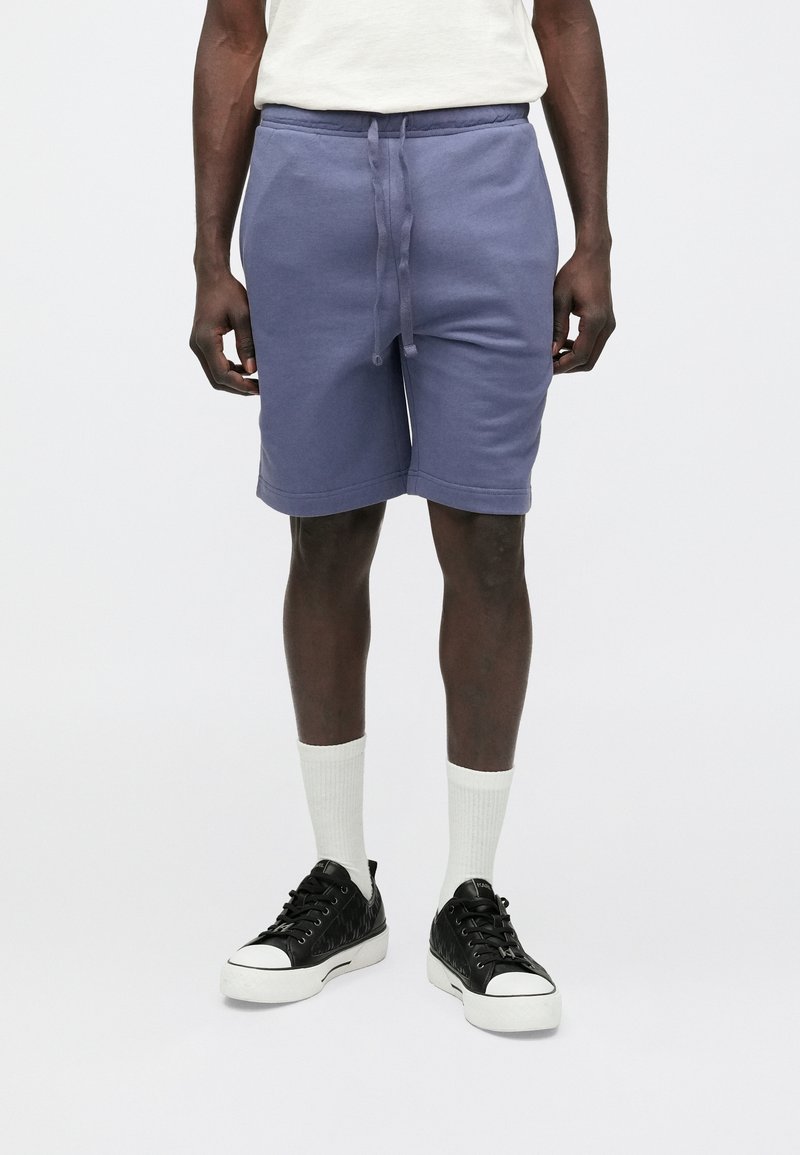 Lilla bomuldsblandede shorts med elastisk talje og snøre, med sidelommer og løs pasform, kombineret med hvide sokker og sorte sneakers.