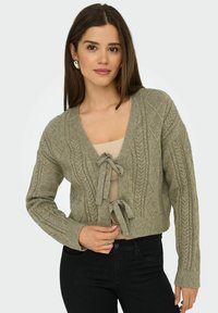 Cardigan en tricot vert olive avec un col en V, présentant un motif en torsades, des liens à l'avant et des poignets côtelés. Porté par-dessus un haut beige et un pantalon noir.