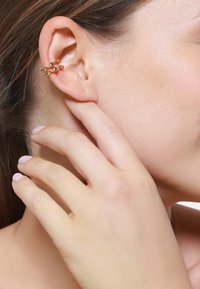 Gouden oorcuff met een gedraaide design, met kleine ronde accenten. De hand van het model raakt voorzichtig haar nek op de achtergrond.