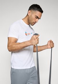 Hvit bomull t-skjorte med flerfarget Nike-logo, kombinert med grå treningsshorts; en person holder treningsmotstandsbånd i begge hender.
