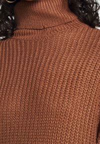 Gros plan d'une personne portant un pull à col roulé en maille côtelée marron, avec des cheveux bouclés foncés visibles sur les bords.