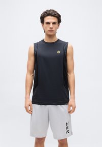 Νέος άνδρας στέκεται με το πρόσωπο προς τα εμπρός φορώντας μαύρο αμάνικο αθλητικό μπλουζάκι και ανοιχτό γκρι αθλητικό σορτς με λογότυπα σε αμφότερα.