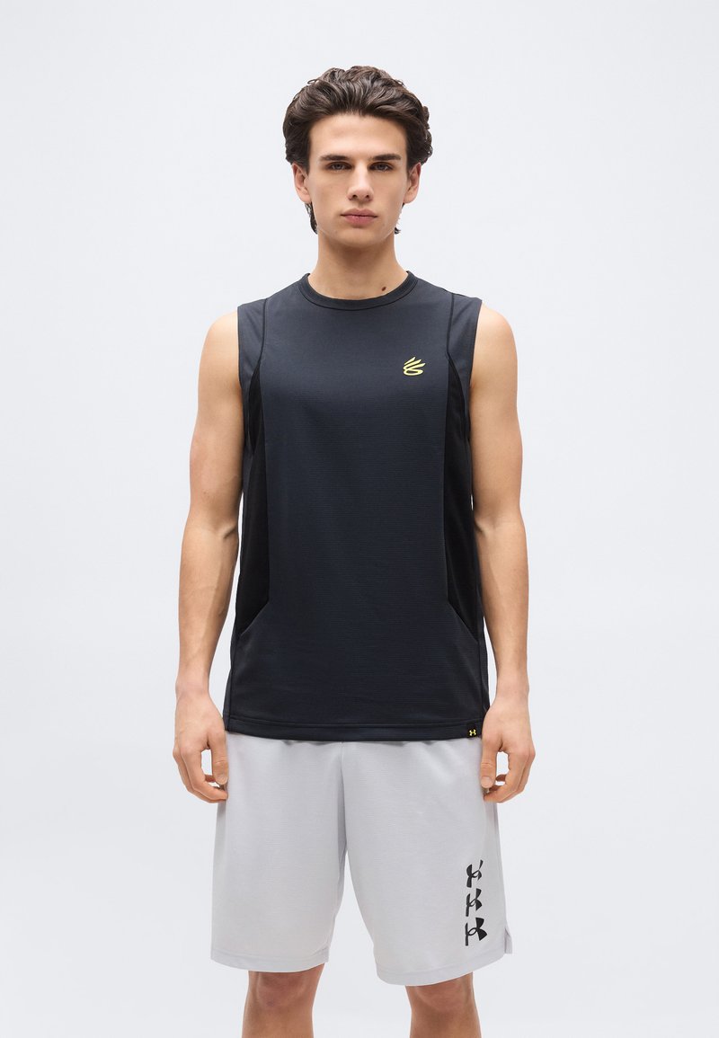 Νέος άνδρας στέκεται με το πρόσωπο προς τα εμπρός φορώντας μαύρο αμάνικο αθλητικό μπλουζάκι και ανοιχτό γκρι αθλητικό σορτς με λογότυπα σε αμφότερα.