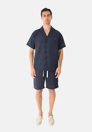 Uomo in piedi che indossa una camicia blu navy a maniche corte con bottoni e pantaloncini coordinati con coulisse, con scarpe beige slip-on su sfondo bianco.