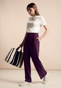 T-shirt blanc, pantalon large violet avec poches, et baskets blanches ; portant un grand sac fourre-tout rayé noir et beige.
