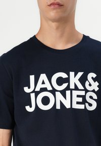Tamsiai mėlynas medvilninis marškinėliai su apykakle, kuriuose ryškiai baltais rašmenimis užrašyta "JACK & JONES" per krūtinę. Trumpomis rankovėmis.